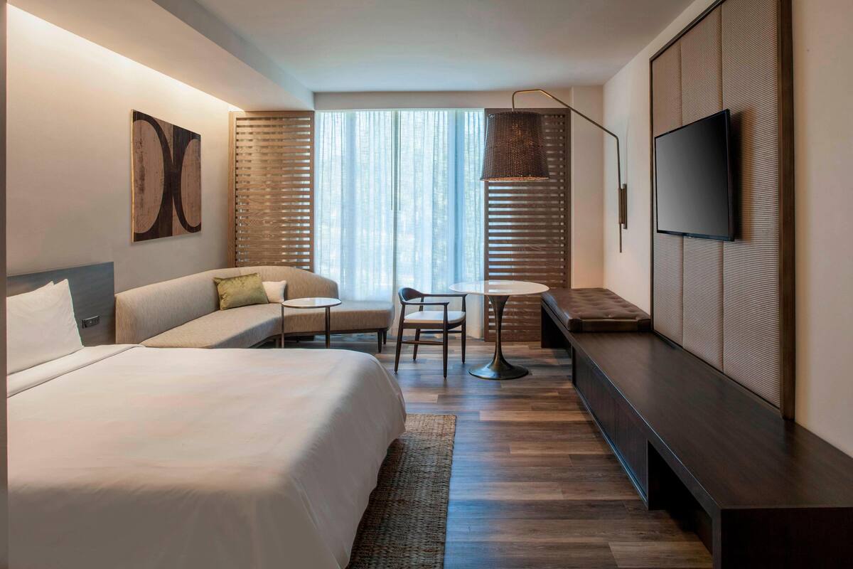 A modern hotel room with a large bed, a sofa, a small round table, and a TV mounted on the wall.