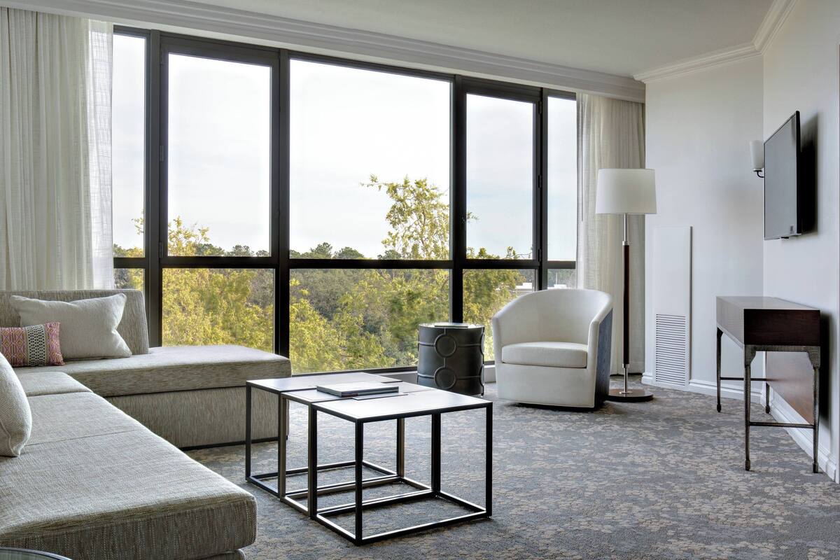A modern hotel room with a large window, a sofa, a coffee table, a chair, a lamp, and a TV.
