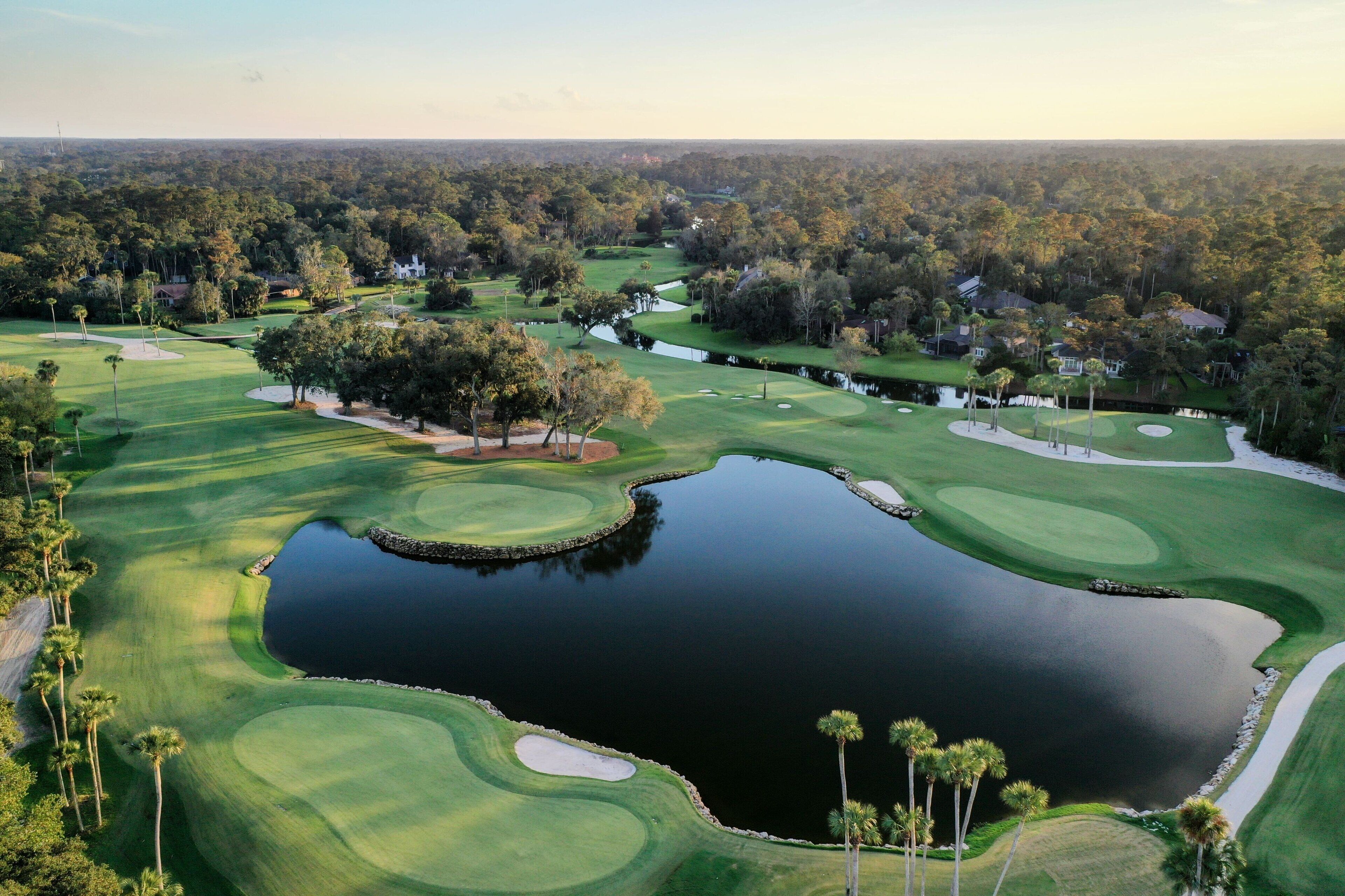 Un campo de golf con un estanque, rodeado de árboles y viviendas.