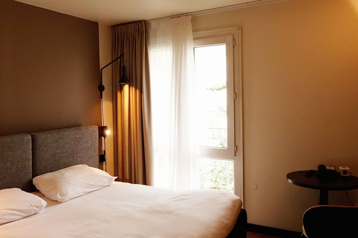 A hotel room with a bed, a desk, a lamp, and a window with curtains.