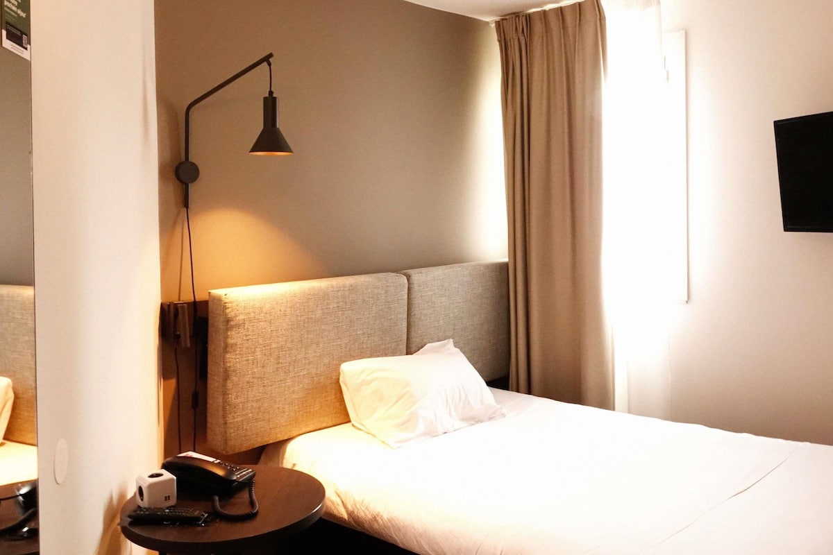 A hotel room with a bed, a bedside table with a lamp, a phone, and a TV mounted on the wall.