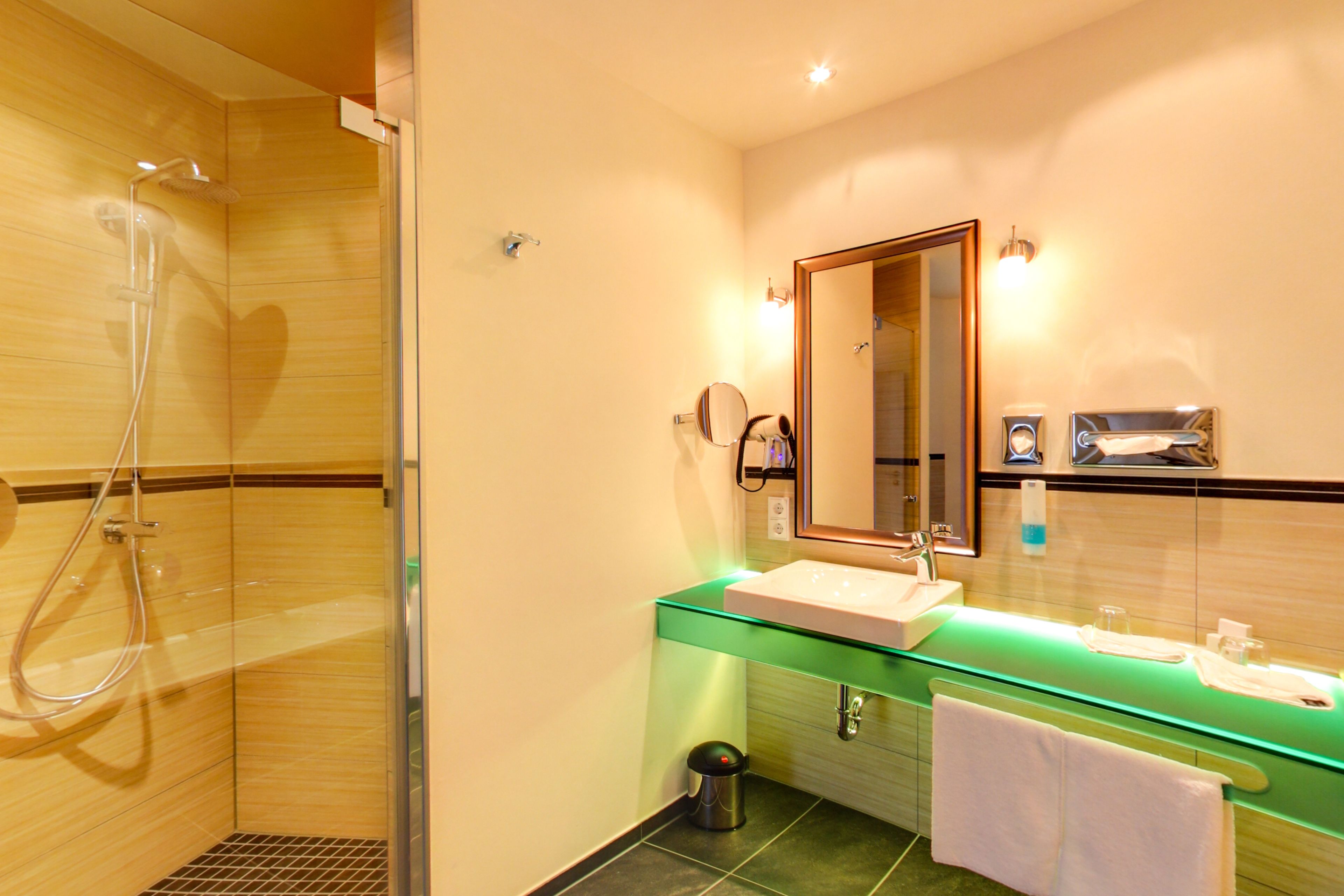 A modern bathroom with a glass shower enclosure, a sink with a green countertop, and a mirror with a wooden frame.