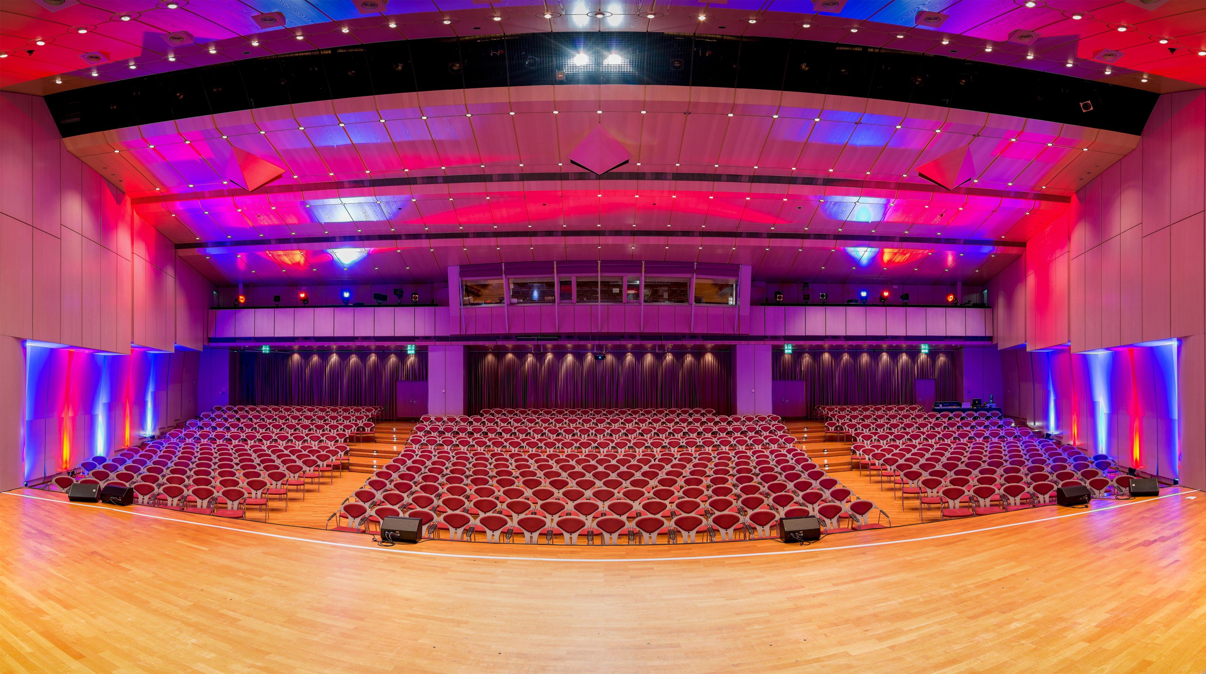A large auditorium with tiered seating, a stage with curtains, and a modern ceiling with lighting fixtures.