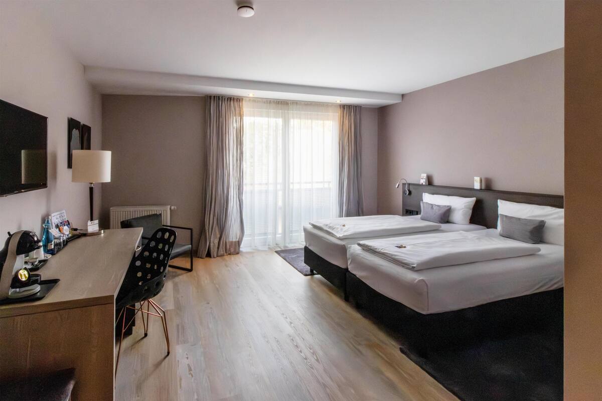 Habitación de hotel con dos camas, un escritorio, una silla, una lámpara y una ventana con cortinas.