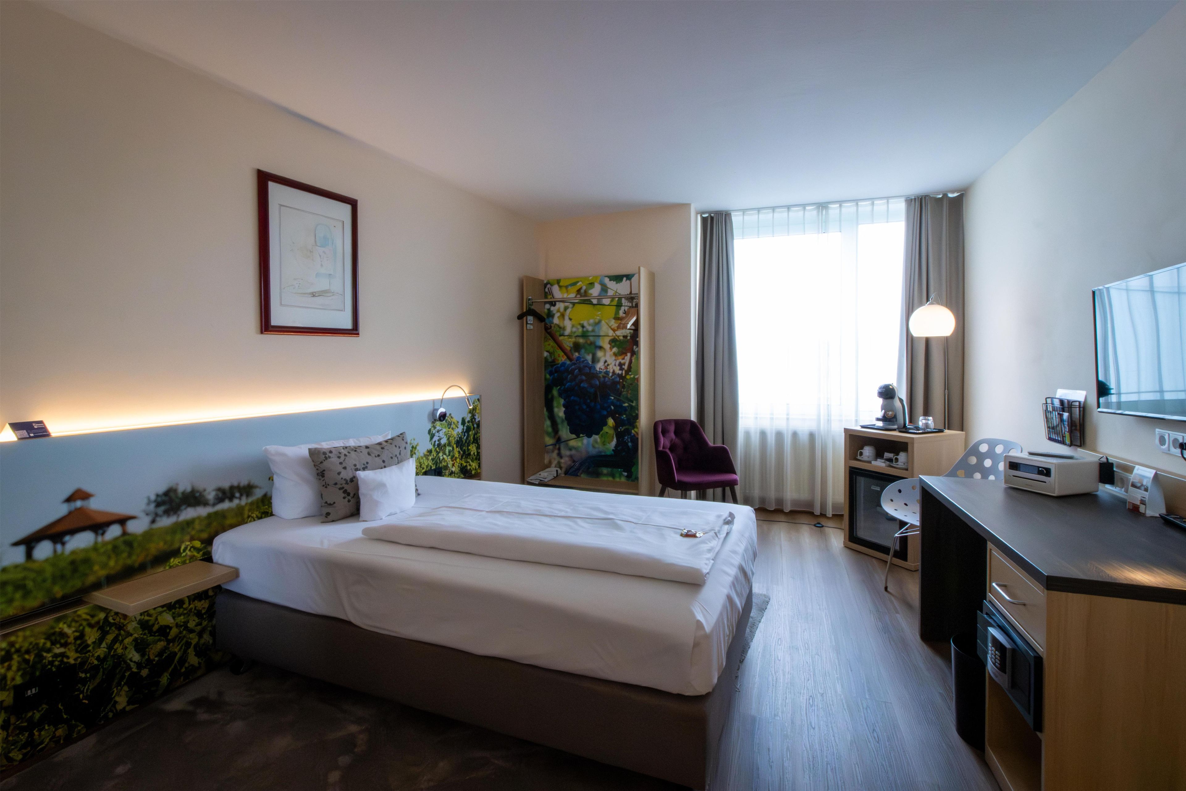 A hotel room with a bed, a desk, a chair, a window with curtains, and a wall-mounted artwork.
