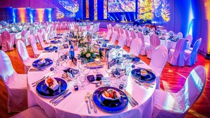 Une salle de banquet avec des tables rondes dressées pour un événement formel, comprenant des chaises blanches, une décoration de table bleue et une scène dotée d’un grand écran.