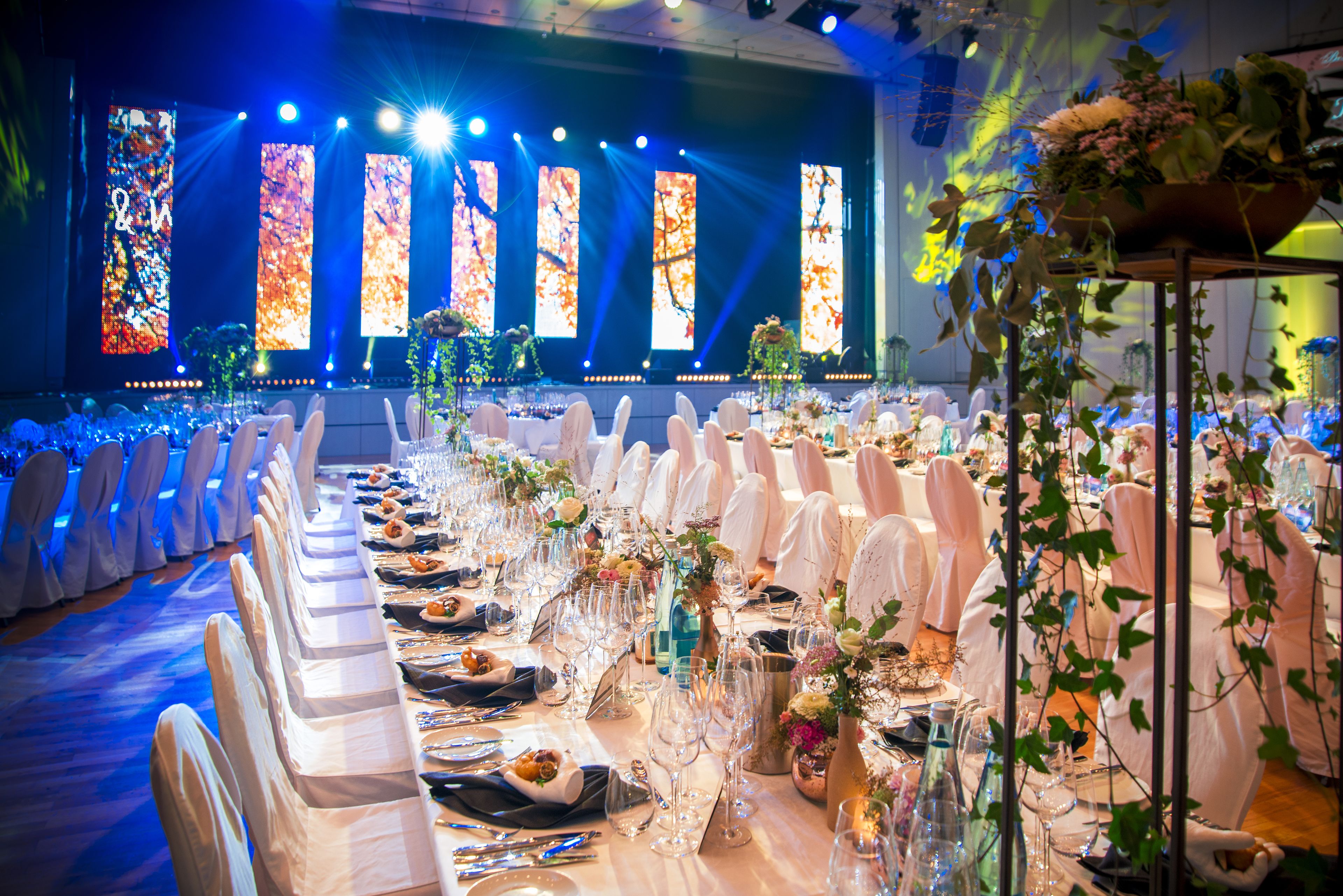 Un salón de banquetes con largas mesas dispuestas para un evento formal, que incluye manteles blancos, sillas y centros de mesa con flores.