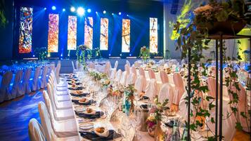Un salón de banquetes con largas mesas dispuestas para un evento formal, que incluye manteles blancos, sillas y centros de mesa con flores.
