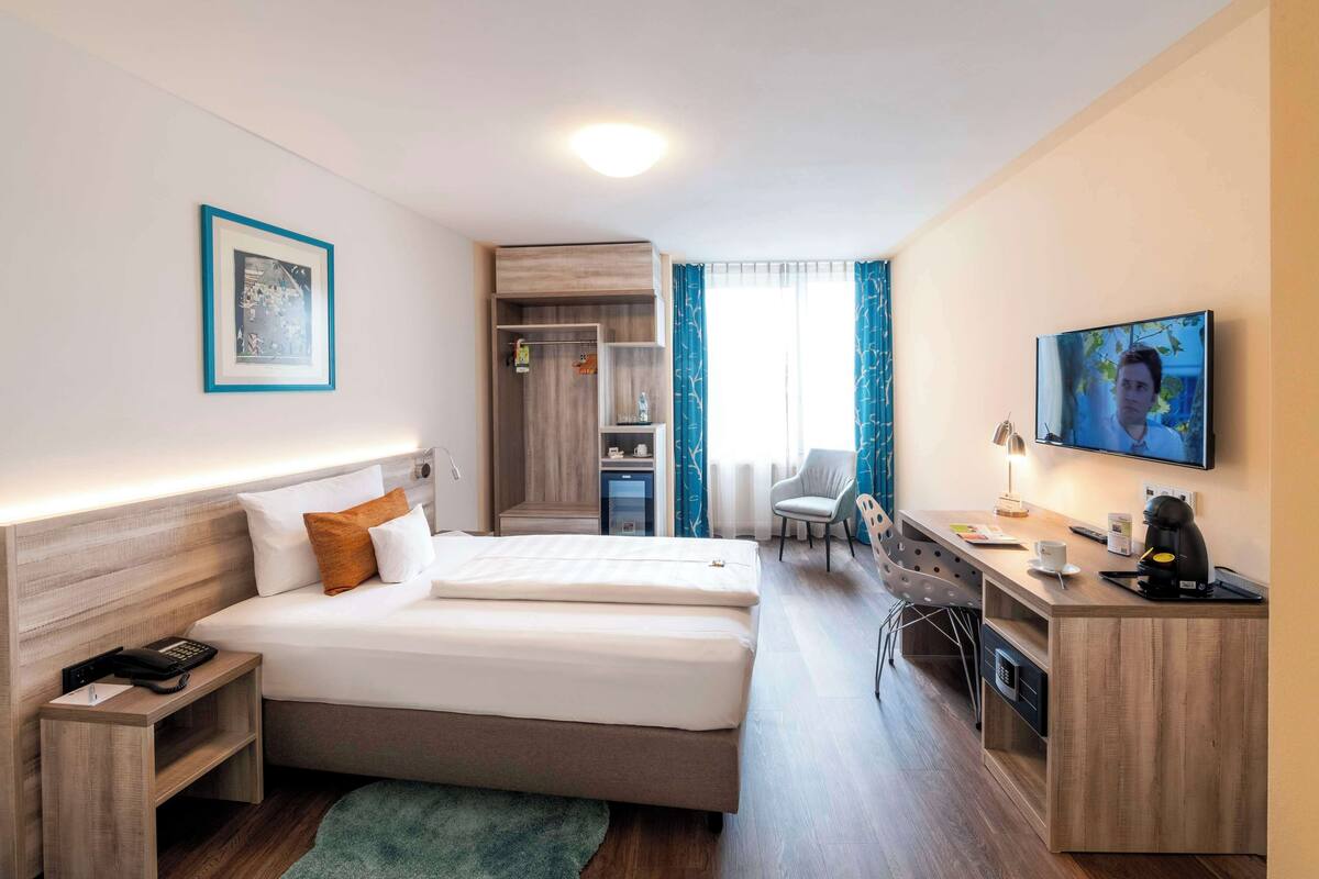 Habitación de hotel con cama, escritorio, silla, televisión y una kitchenette pequeña.