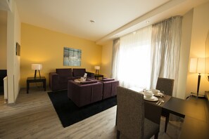 A hotel room with a sofa, a dining table set for two, a painting on the wall, and a large window with curtains. - Best Western Plus Palatin Kongresshotel (Wiesloch)
