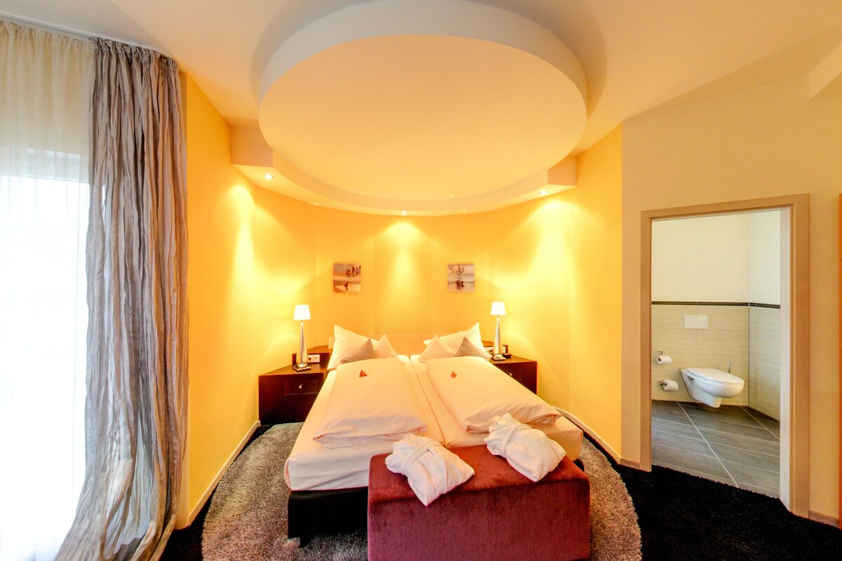 Habitación de hotel con cama doble, dos mesitas de noche con lámparas, un taburete rojo y un baño con inodoro y lavabo.