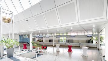 A spacious lobby with a modern design, featuring a seating area with red and white chairs, a reception desk, and a large glass wall offering a view of the outdoors.