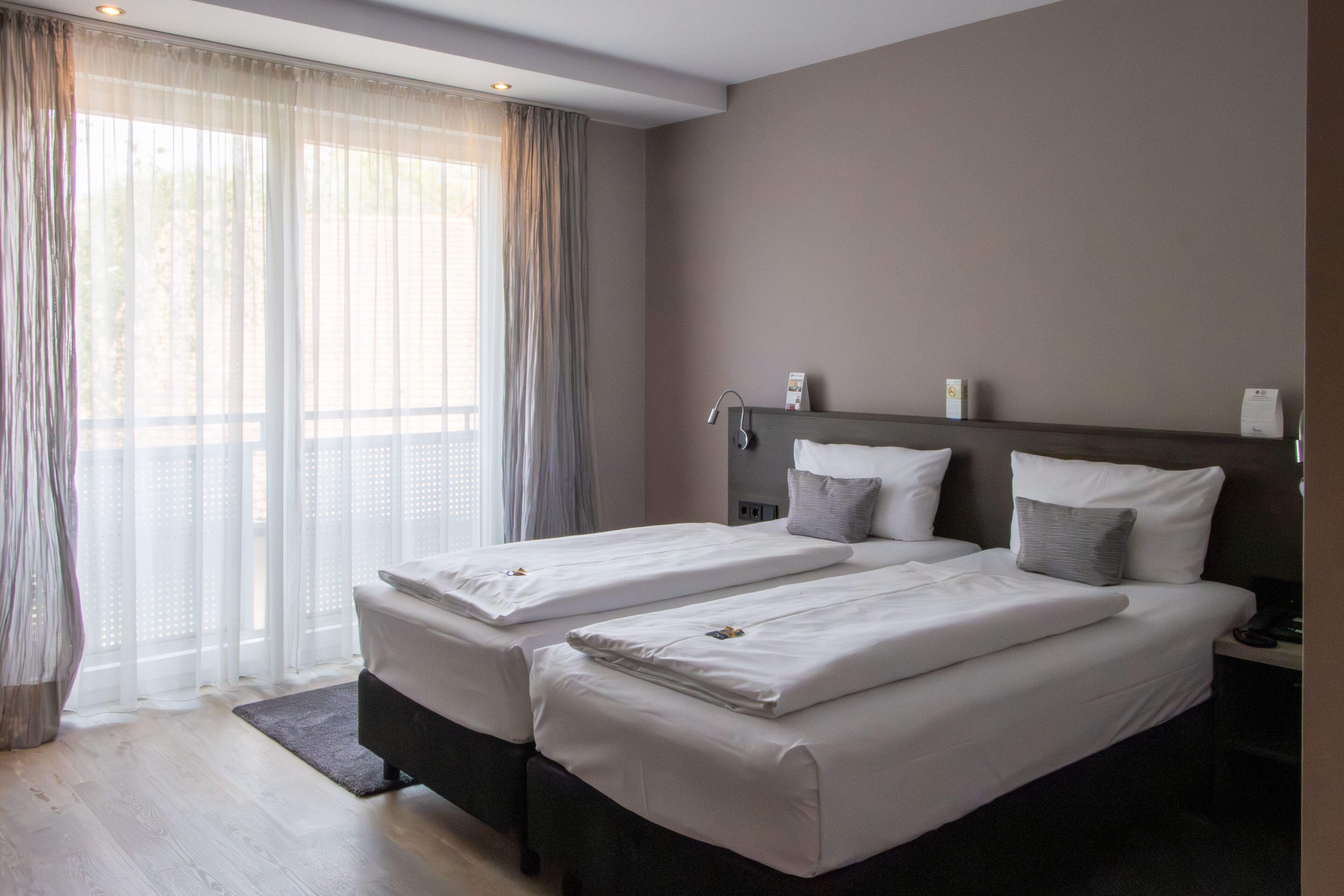A hotel room with two beds, a large window with sheer curtains, a nightstand with a lamp, and a small table with a phone.