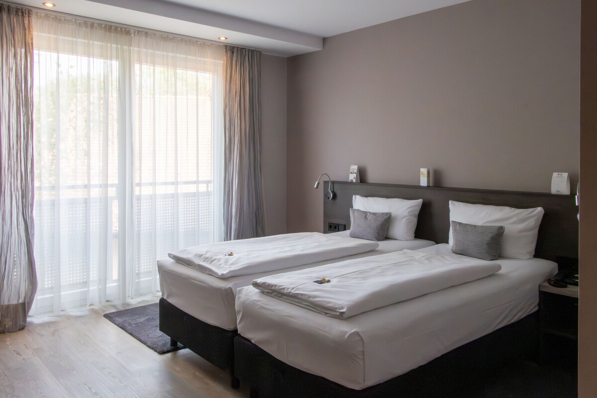 Una habitación de hotel con dos camas, un ventanal con cortinas translúcidas, una mesita de noche con lámpara y una pequeña mesa con teléfono.