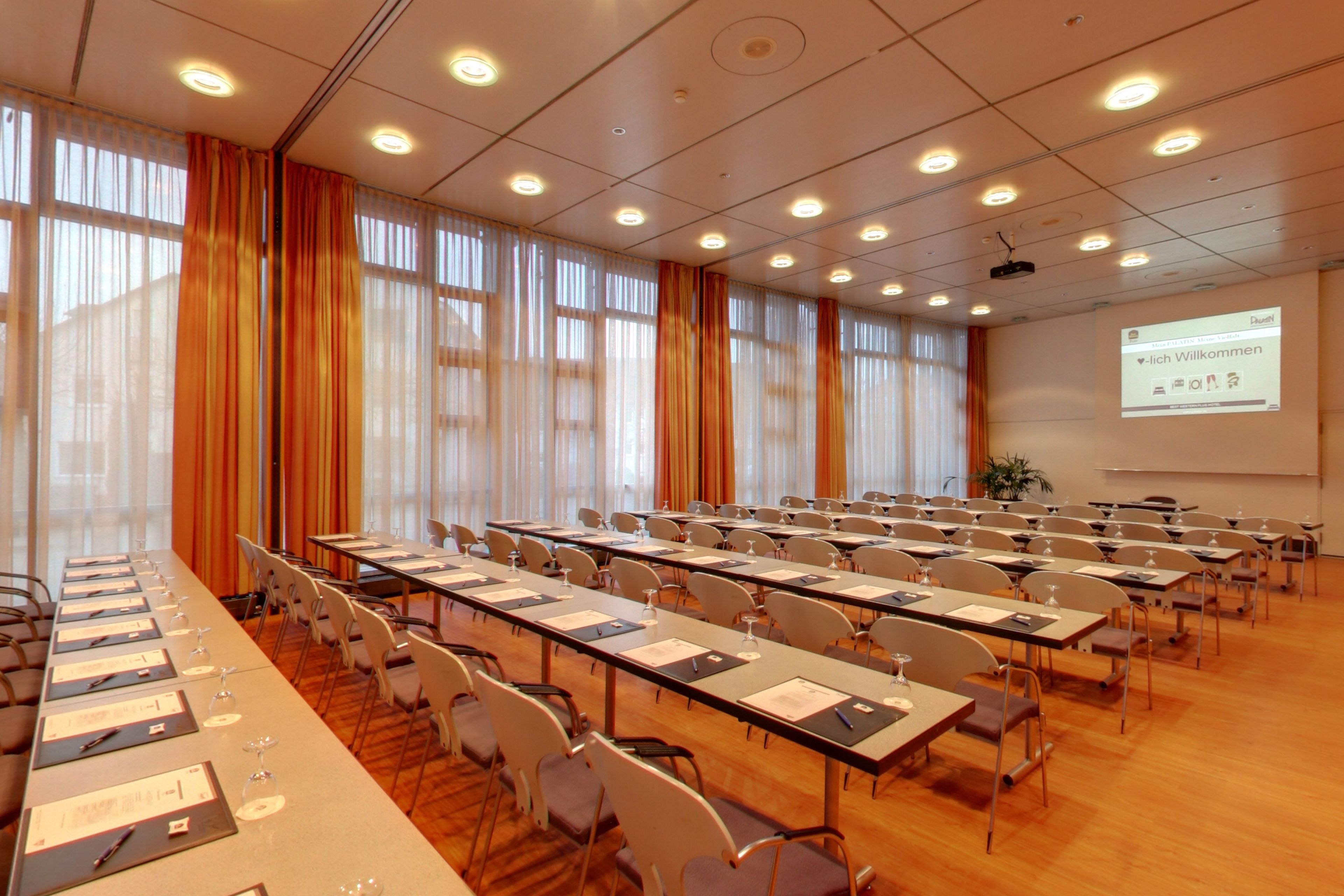 Een congreszaal met rijen tafels, stoelen en een projectiescherm waarop een welkomstboodschap staat.