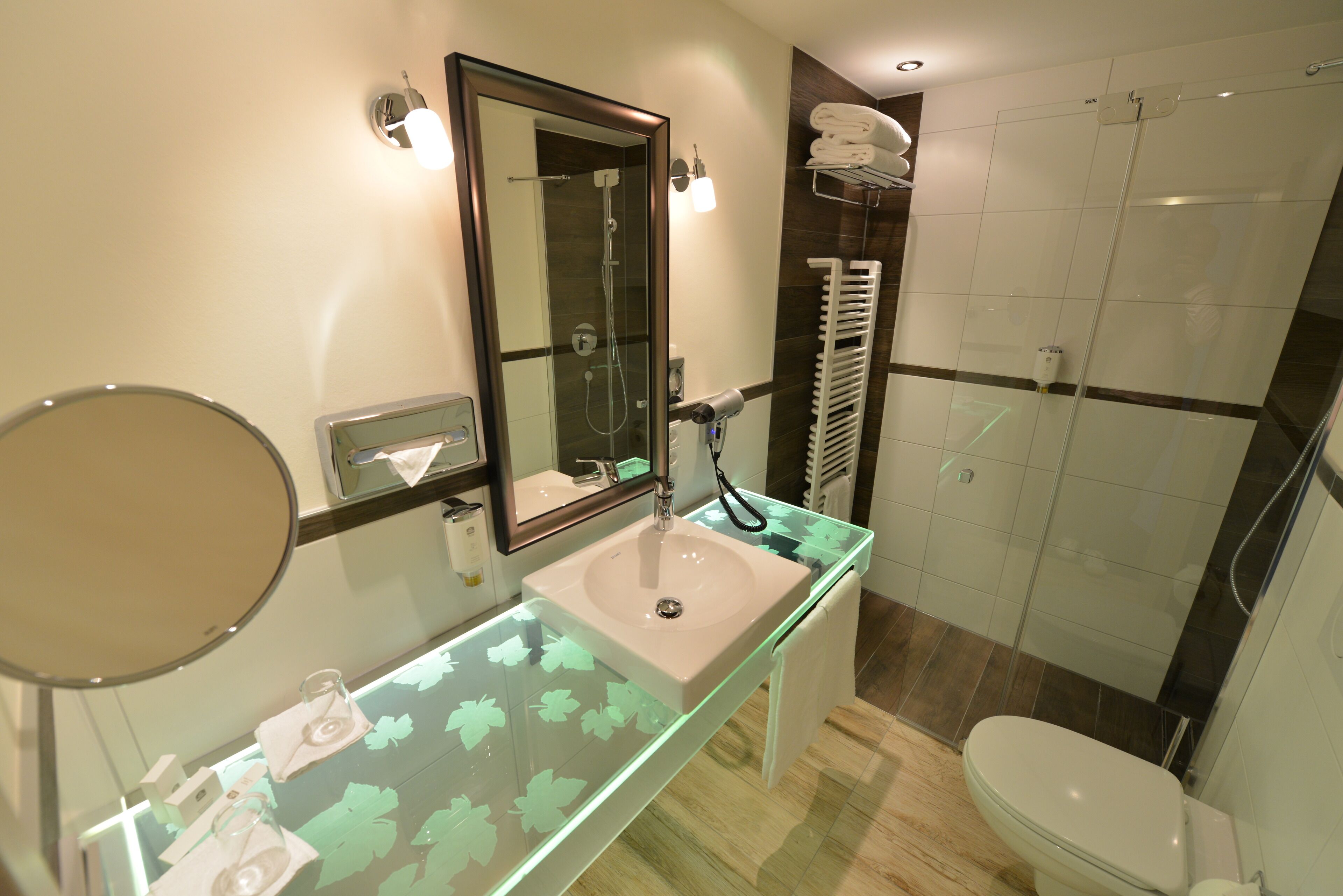 A modern bathroom with a glass vanity, a large mirror, a shower enclosure, and a toilet.