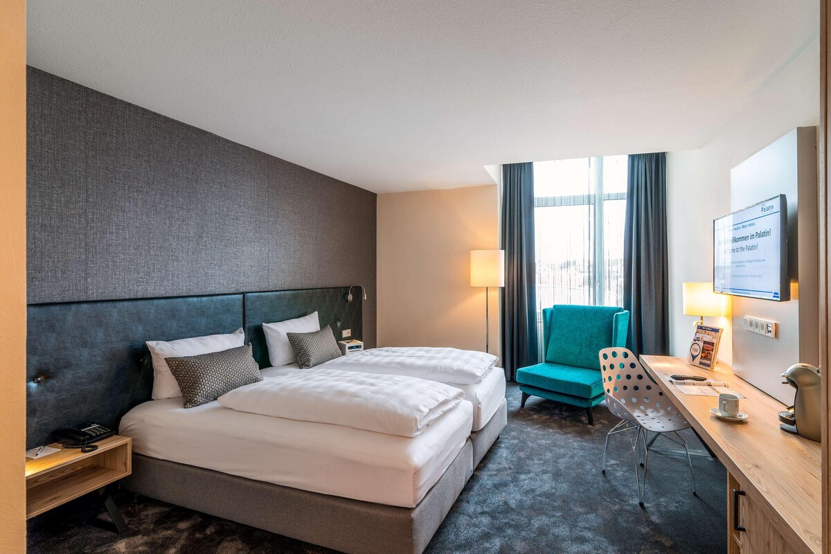 Une chambre d’hôtel avec un lit, un bureau, une chaise, une lampe et une fenêtre avec des rideaux.