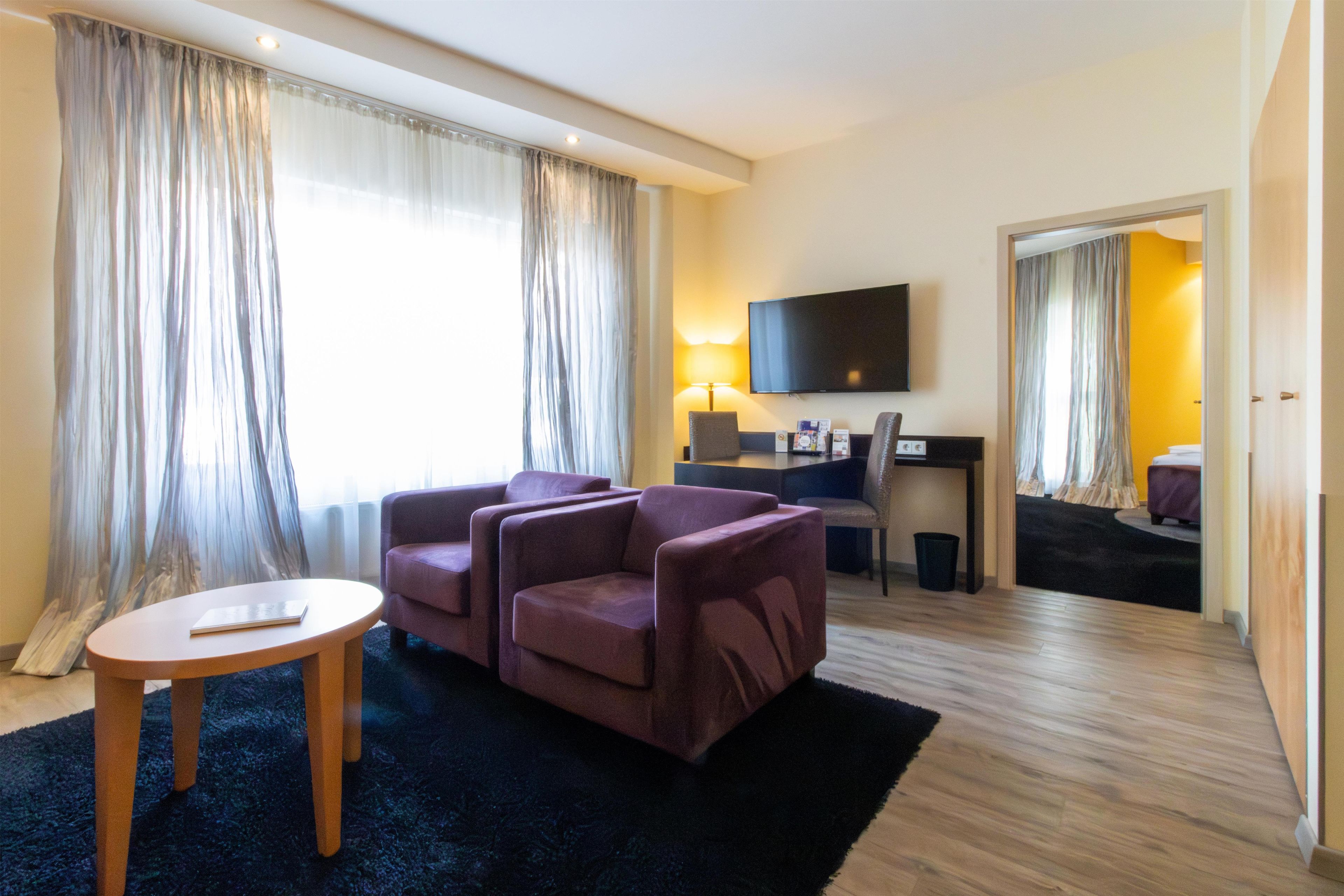 A hotel room with a seating area, a flat-screen TV, a small table, and a view of the adjacent room.