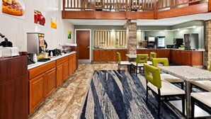 A hotel kitchen with wooden cabinets, a dining area with green chairs, a kitchen island, and a view of the kitchen from the dining area. - Quality Inn Mason Near Kings Island (Mason)