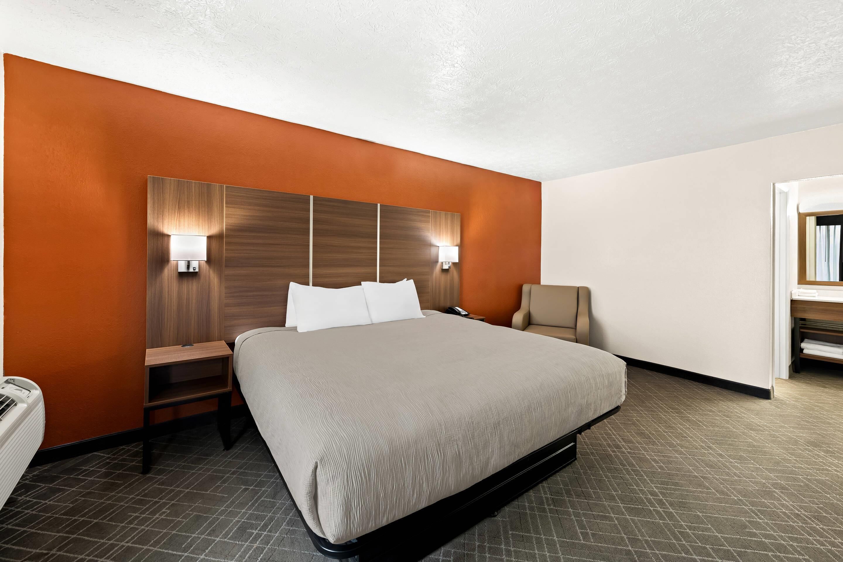 A hotel room with a large bed, a nightstand, a chair, and a desk with a lamp.