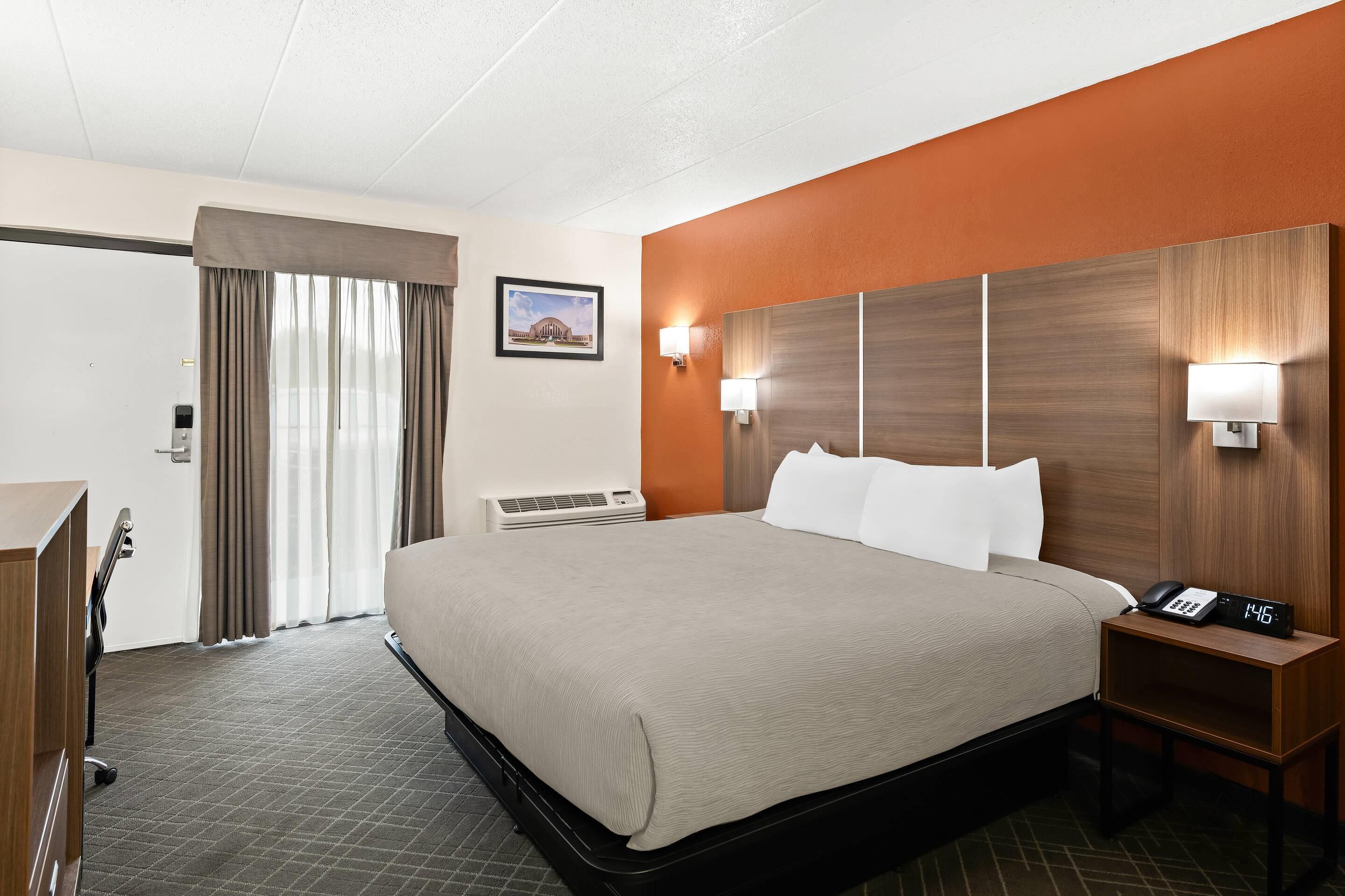 A hotel room with a large bed, a desk, a chair, a nightstand with a phone and clock, and a picture on the wall.