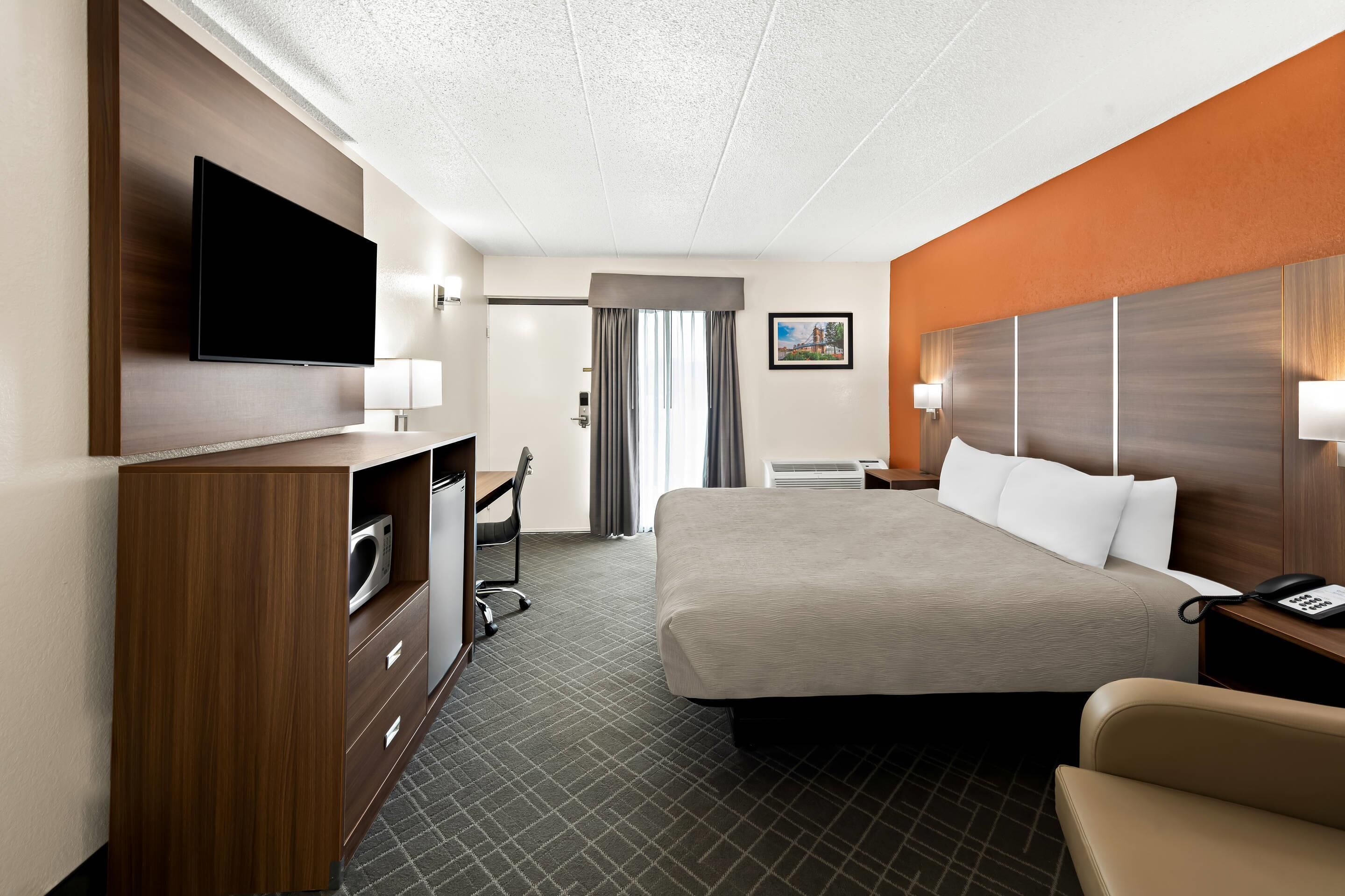 A hotel room with a large bed, a desk with a flat-screen TV, a chair, a desk lamp, and a nightstand with a phone.