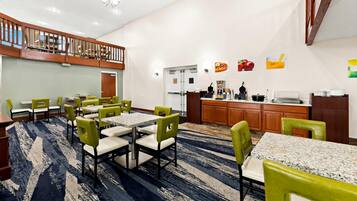 A hotel dining area with multiple tables and chairs, a kitchen in the background, and a balcony with wooden railings.