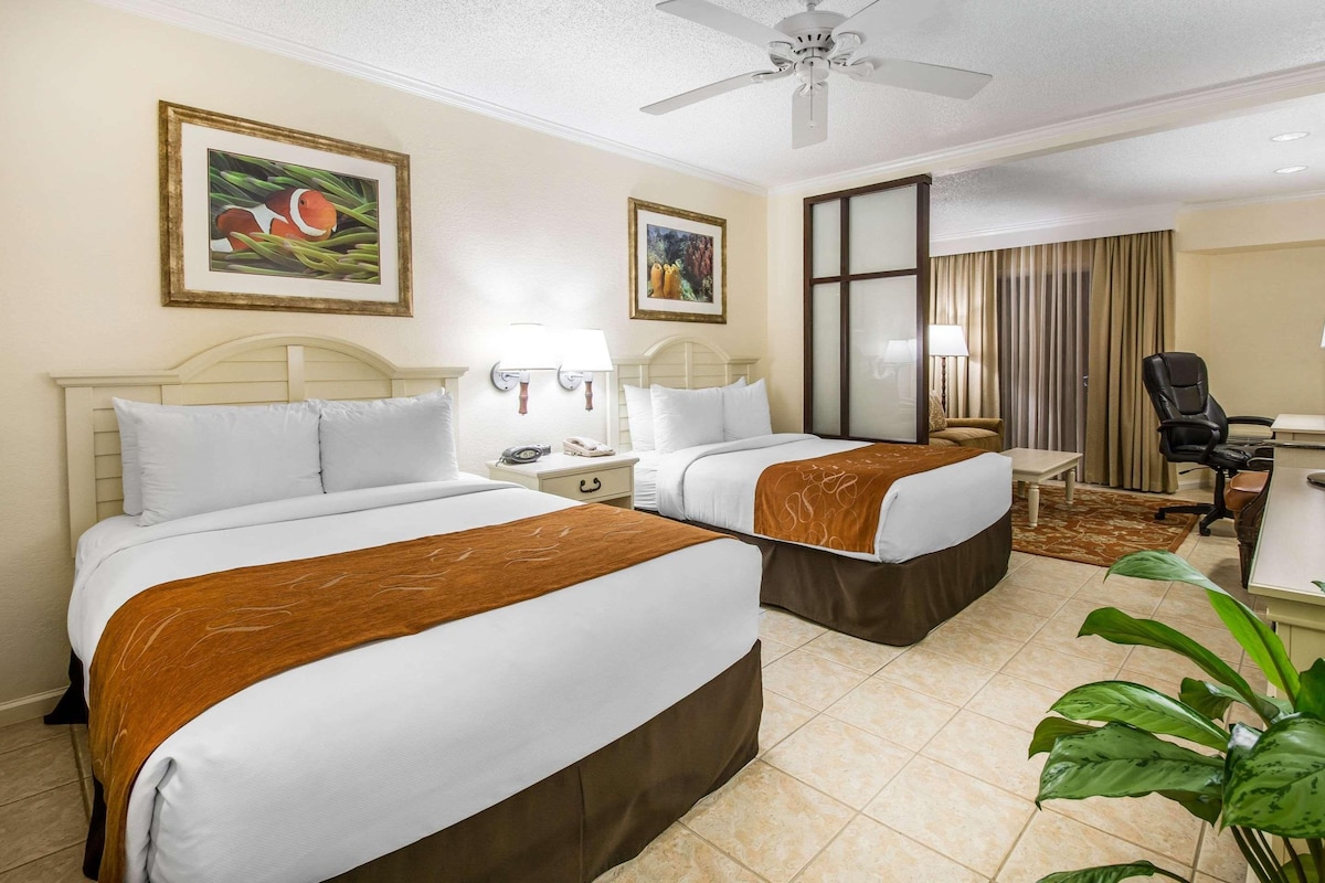 A hotel room with two beds, a desk, a chair, a ceiling fan, and two framed pictures on the wall.
