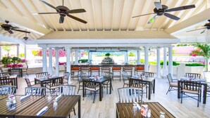 An outdoor dining area with multiple tables and chairs, ceiling fans, and a bar in the background.