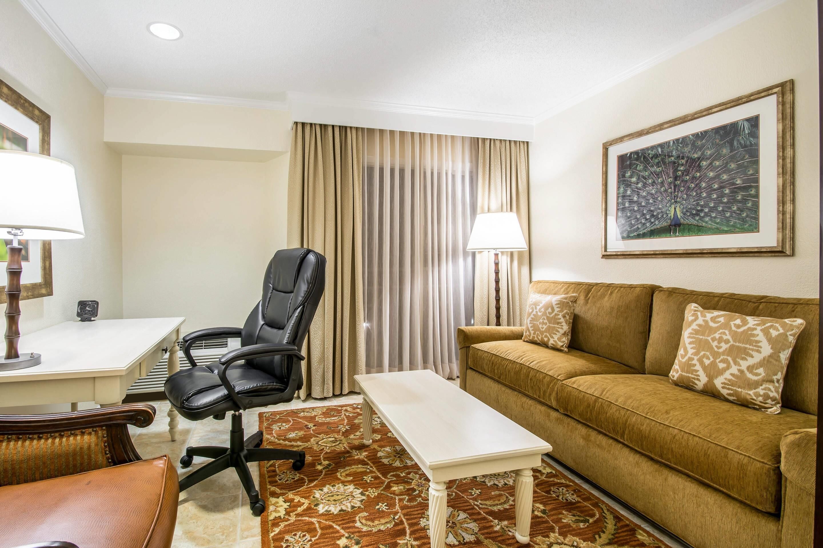 Quarto de hotel com escrivaninha, cadeira de escritório preta, sofá marrom com almofadas estampadas, mesa de centro branca, tapete estampado, uma grande moldura na parede e janela com cortinas bege.