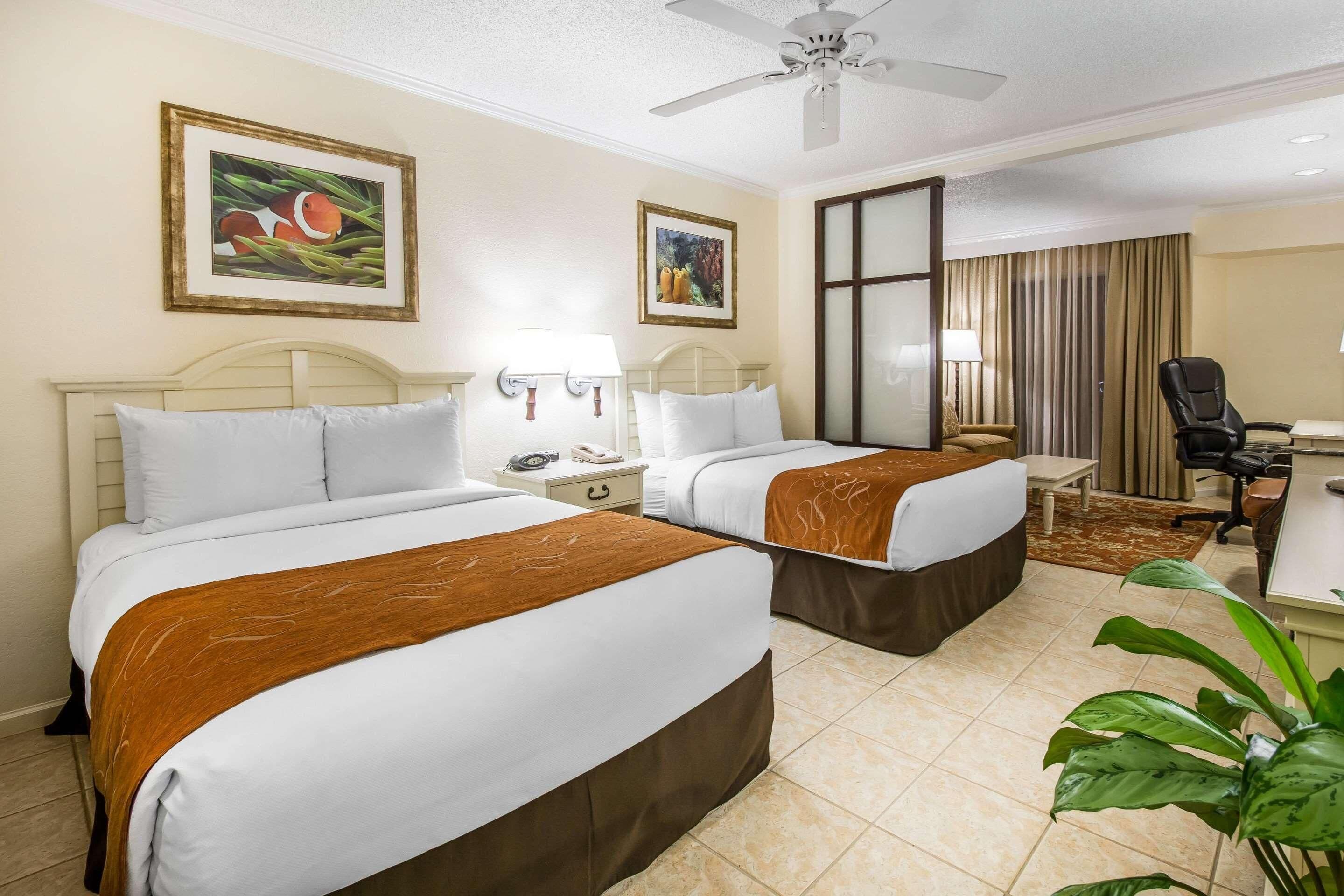 A hotel room with two beds, a desk, a chair, a ceiling fan, and two framed pictures on the wall.