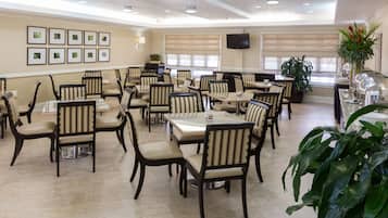 A spacious dining area with multiple tables and chairs, a flat-screen TV mounted on the wall, and several potted plants.