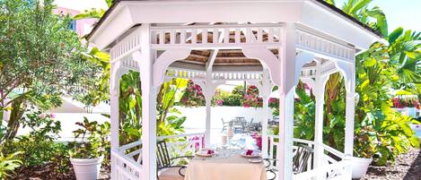 A white gazebo with a table set for two, surrounded by lush tropical plants.