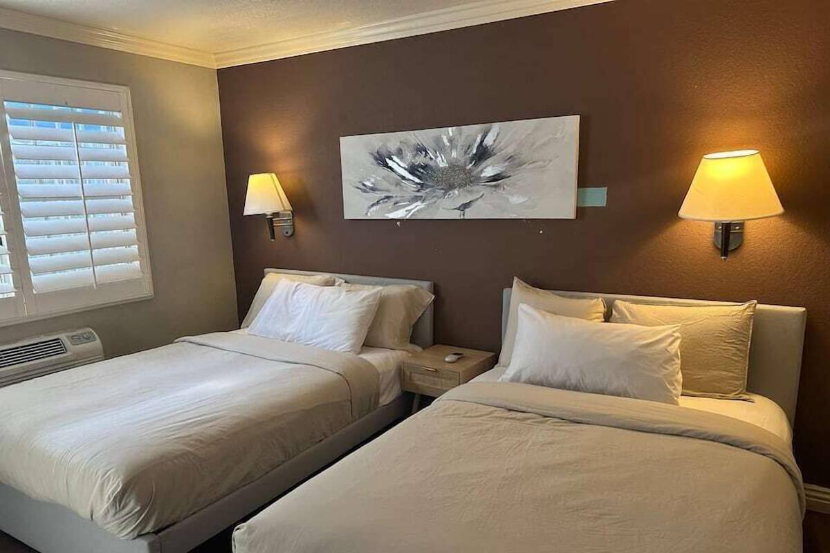 A hotel room with two beds, a window with blinds, a wall-mounted abstract painting, and two wall lamps.