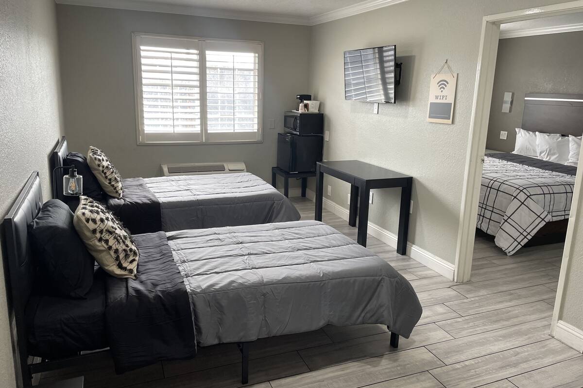 A bedroom with two beds, a small table, a TV, and a window with blinds.