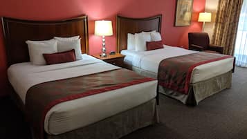 Habitación de hotel con dos camas, cada una con cabecero de madera, ropa de cama blanca y almohadas decorativas rojas. Hay una mesita de noche con lámpara, una silla y un cuadro en la pared.