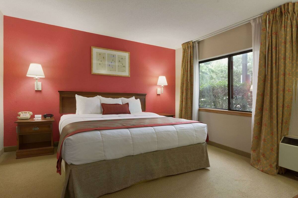 A hotel room with a bed, two bedside lamps, a telephone, a framed picture on the wall, and a window with curtains.