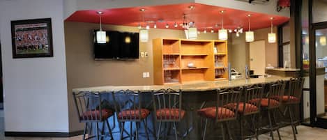 A bar area with multiple stools, a television mounted on the wall, and shelves with glasses.