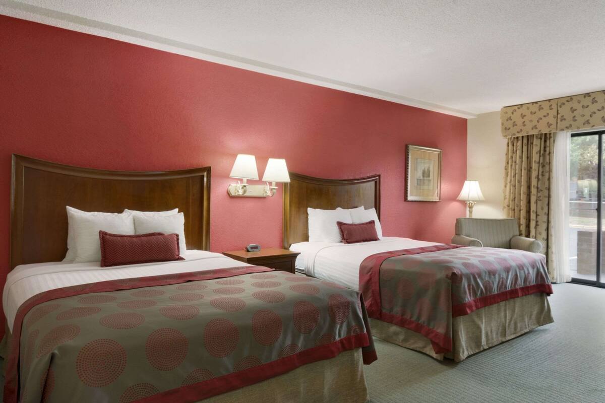 A hotel room with two beds, a nightstand, a lamp, and a window with curtains.