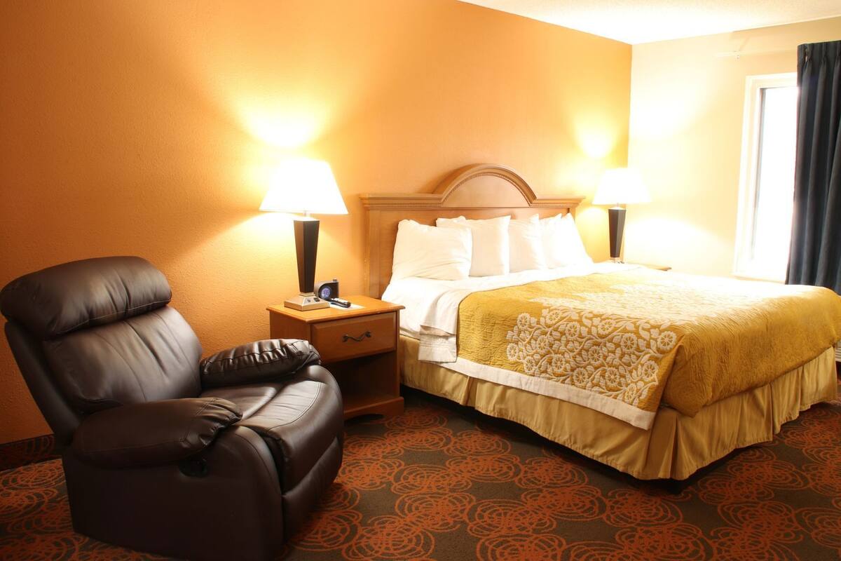 Ein Hotelzimmer mit Ledersessel, Bett mit gemustertem Überwurf, zwei Nachttischlampen und einem Nachttisch mit Fernbedienung.