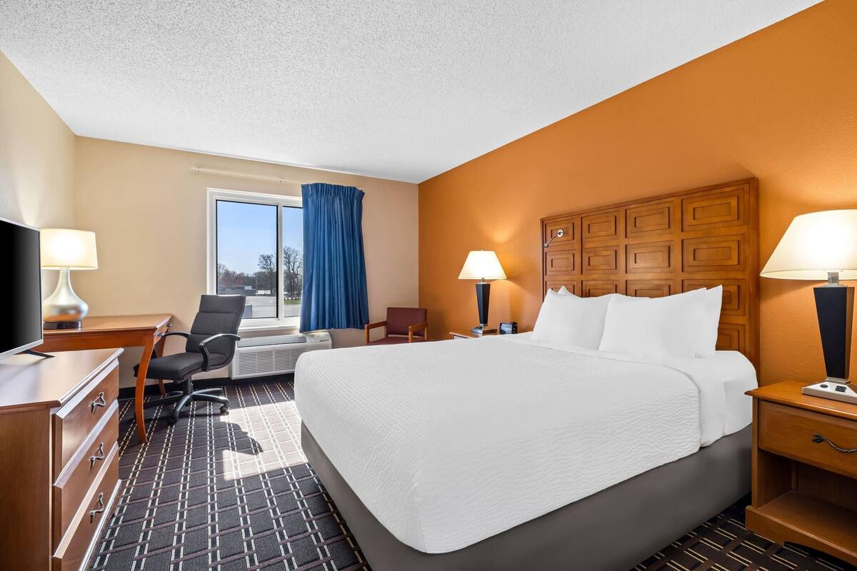 Ein Hotelzimmer mit einem großen Bett, einem Schreibtisch mit Computer, einem Sessel, einem Fernseher und einem Fenster mit Vorhängen.
