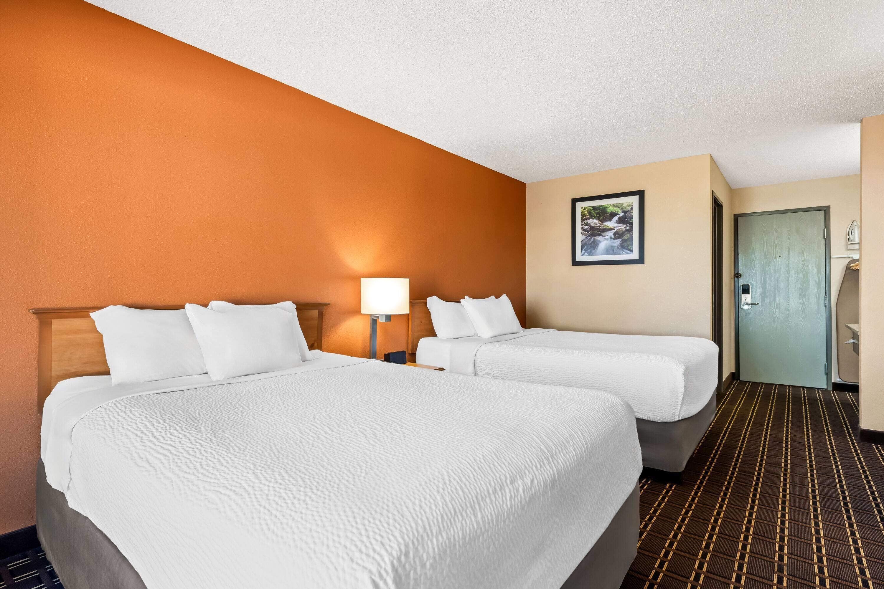 A hotel room with two beds, a nightstand with a lamp, a framed picture on the wall, and a door leading to another room.