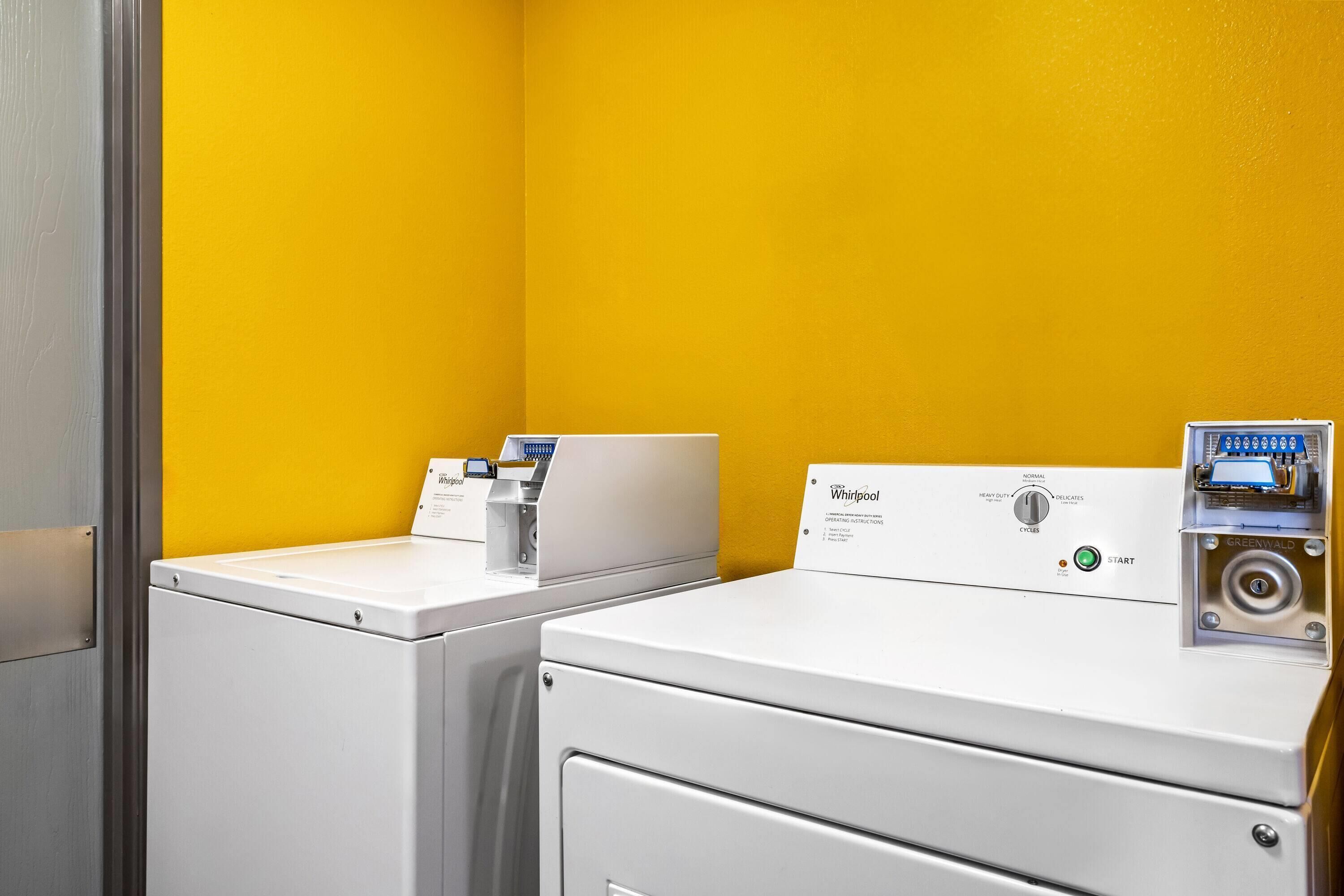 Een wasserette met twee witte wasmachines en drogers tegen een gele muur.
