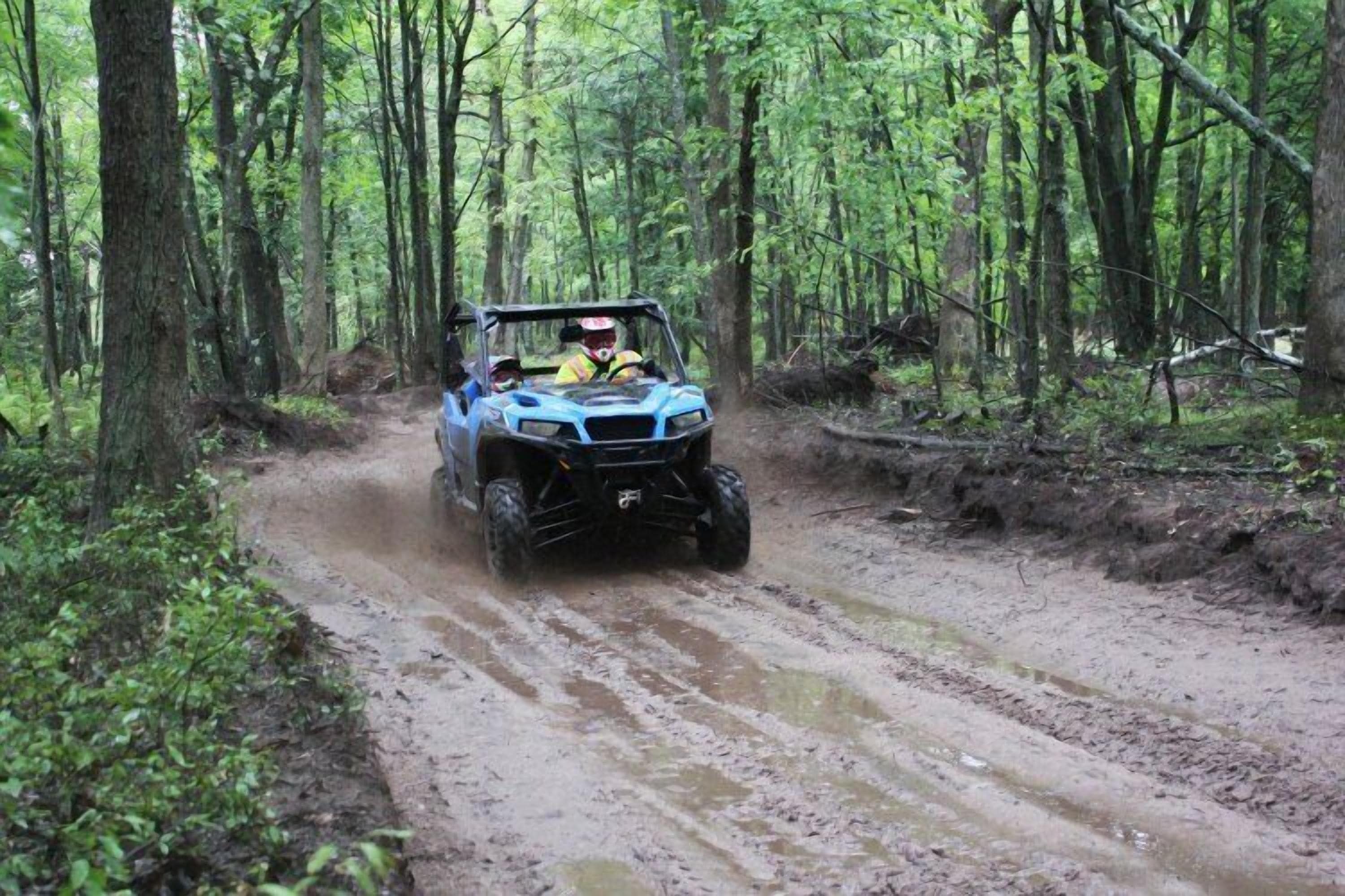 A blue off-road vehicle driving on a muddy path through a forest.