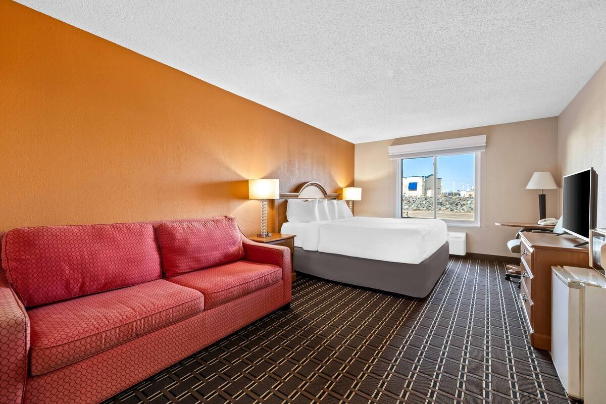 Ein Hotelzimmer mit einem roten Sofa, einem Bett, einem Schreibtisch mit Fernseher und einem Fenster mit Blick auf Gebäude.