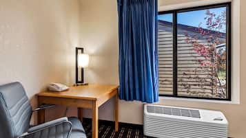 Habitación de hotel con escritorio, silla, aire acondicionado y ventana con vista a un edificio y un árbol.