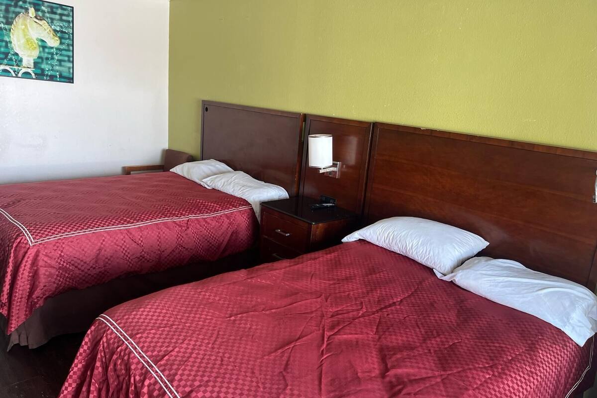 A hotel room with two single beds, each with a red quilt and white pillows. There is a wooden headboard, a nightstand with a lamp, and a wall-mounted artwork of a horse.