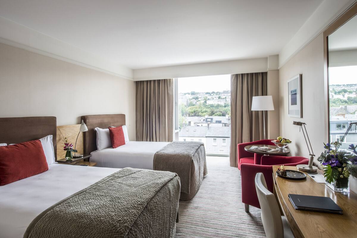 Une chambre d’hôtel avec deux lits, un bureau, une chaise, une lampe, un vase avec des fleurs et une vue sur la ville.