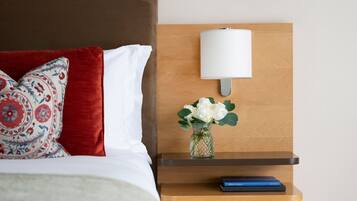 Una cama bien hecha con un cojín rojo estampado, un cojín blanco y un florero con flores blancas sobre una mesita de noche de madera.