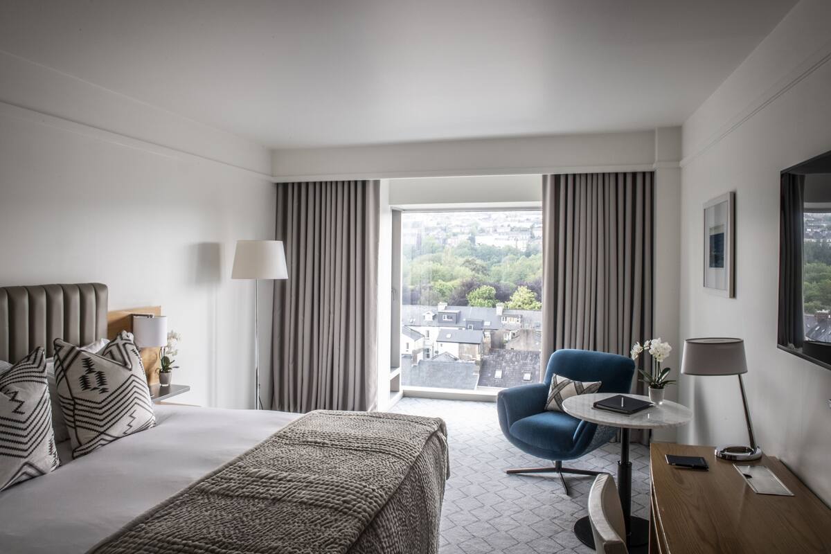 Une chambre d’hôtel avec un lit, un bureau, une chaise, une lampe et une vue sur la ville par la fenêtre.